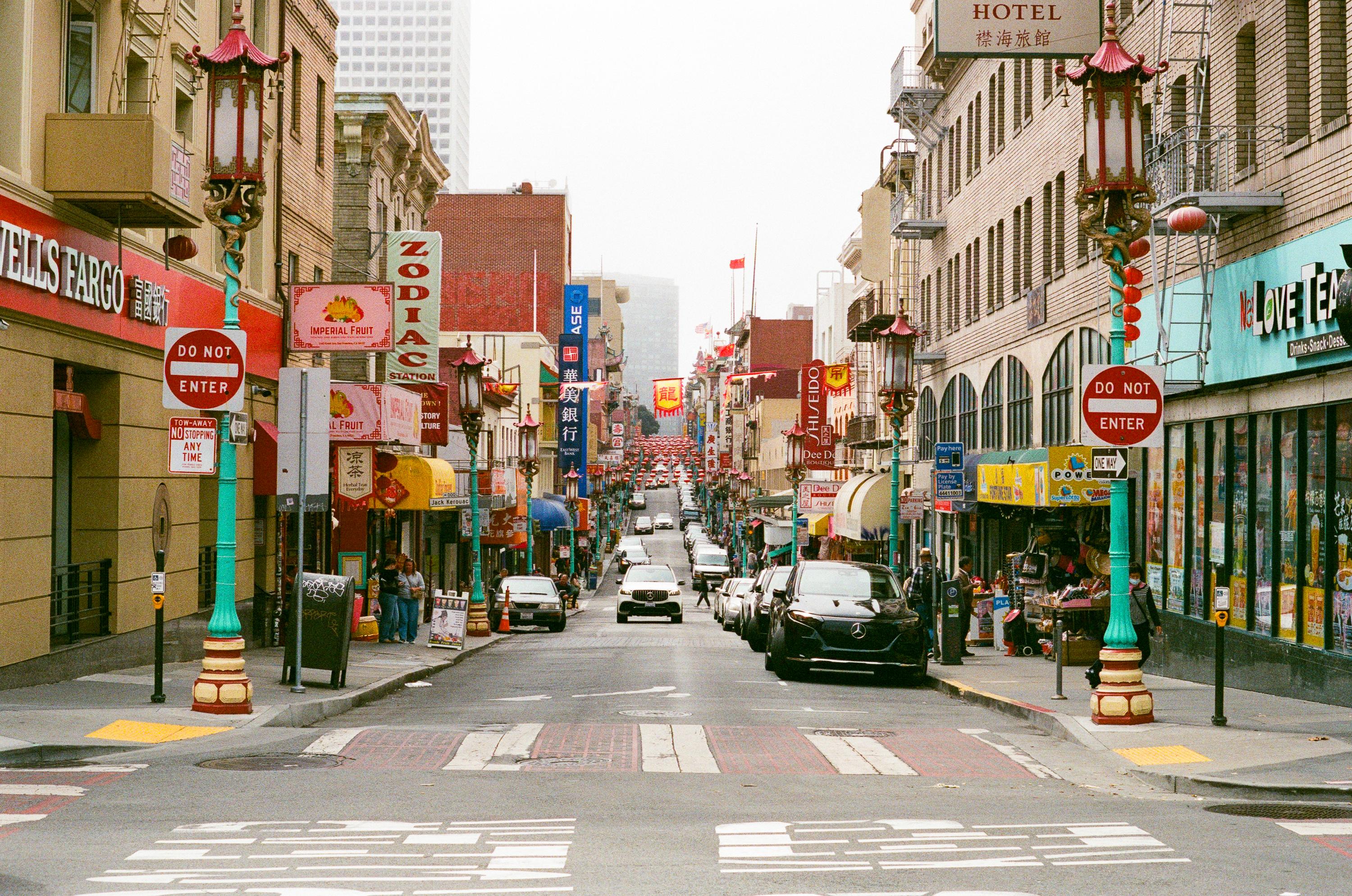 Busy urban street lined with shops, restaurants, and colorful signs, featuring pedestrians, parked cars, and decorative street lamps with red lanterns.