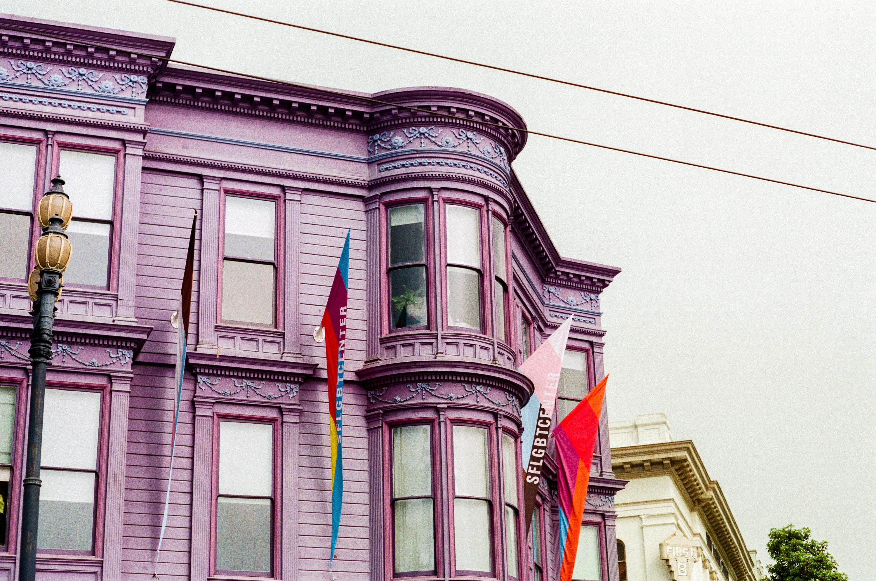 Purple Victorian-style building with ornate details, bay windows, and rainbow flags hanging outside on a cloudy day.