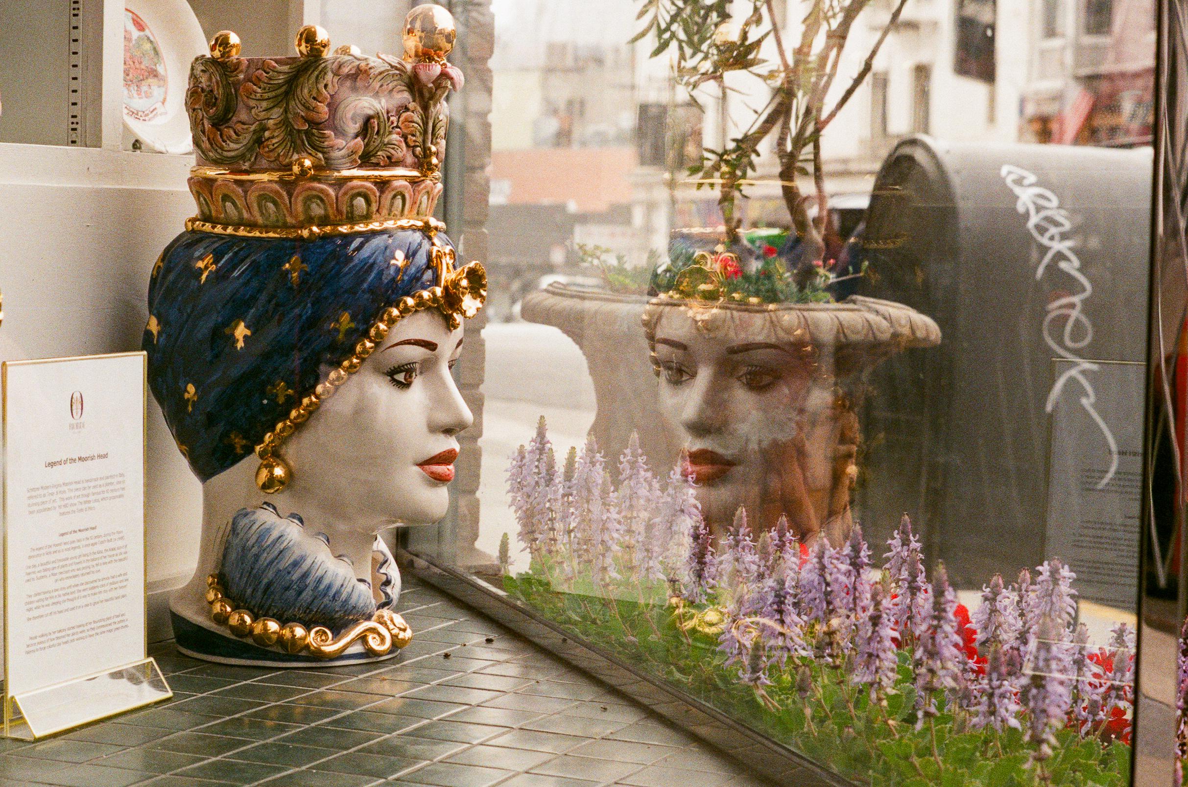 Decorative ceramic head with a crown and jewelry displayed by a window, reflecting in the glass with purple flowers and urban street scene outside.