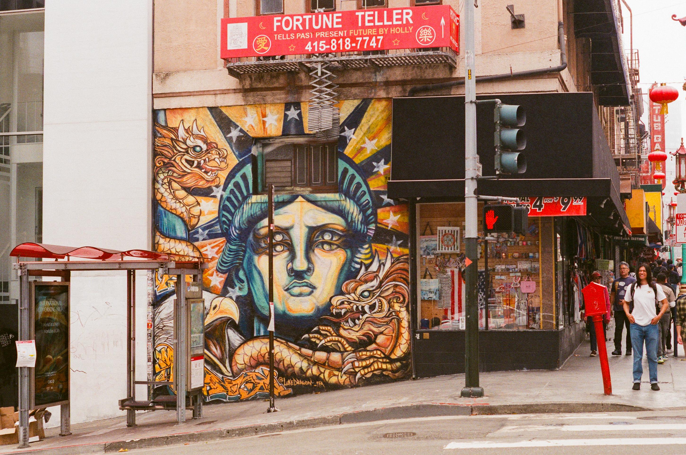 Street corner with colorful mural of Statue of Liberty and dragons, a fortune teller sign above, and pedestrians walking nearby in an urban setting.