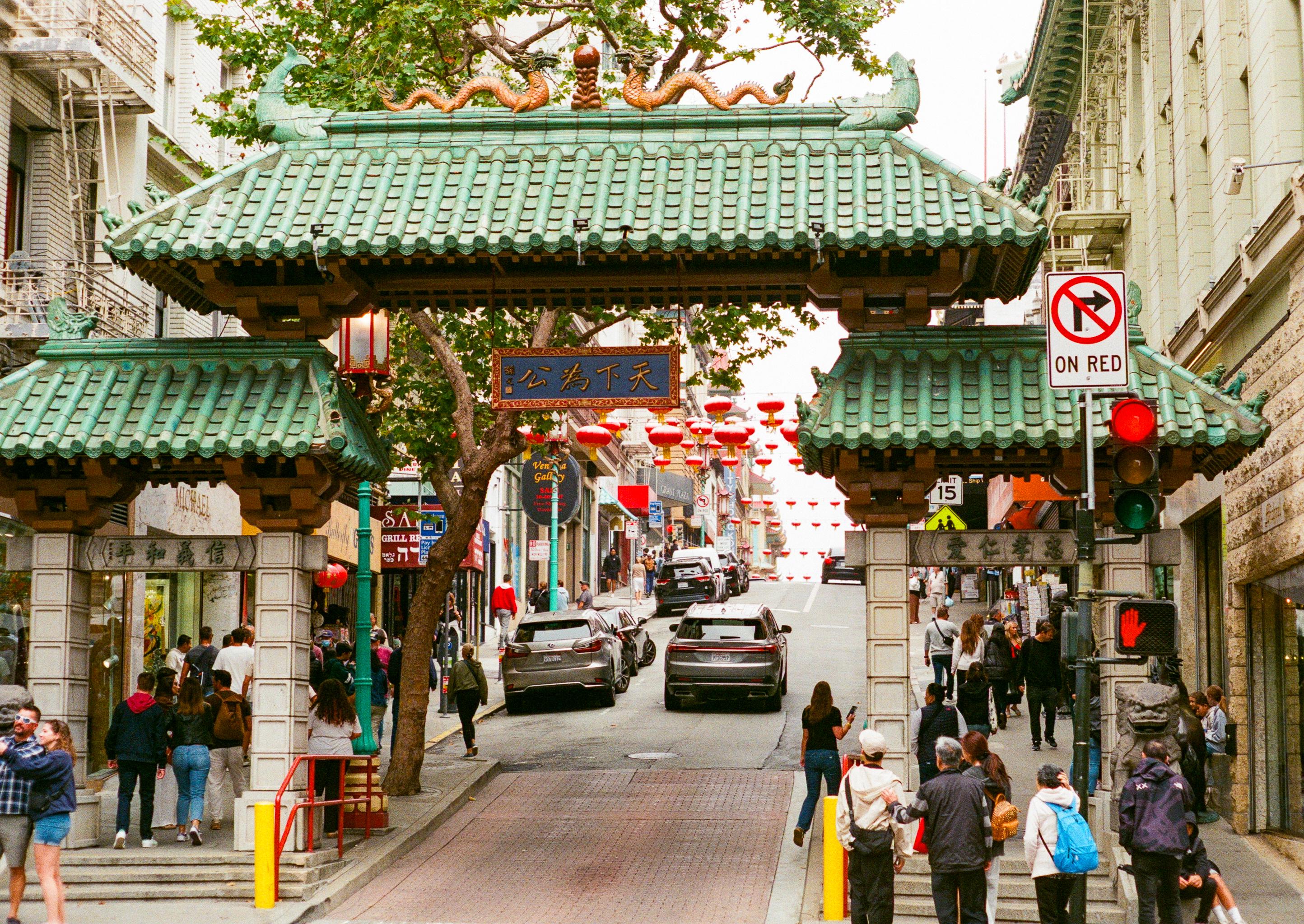 Entrance gate to Chinatown with green tiled roof, red lanterns, and people walking on the street in an urban setting.
