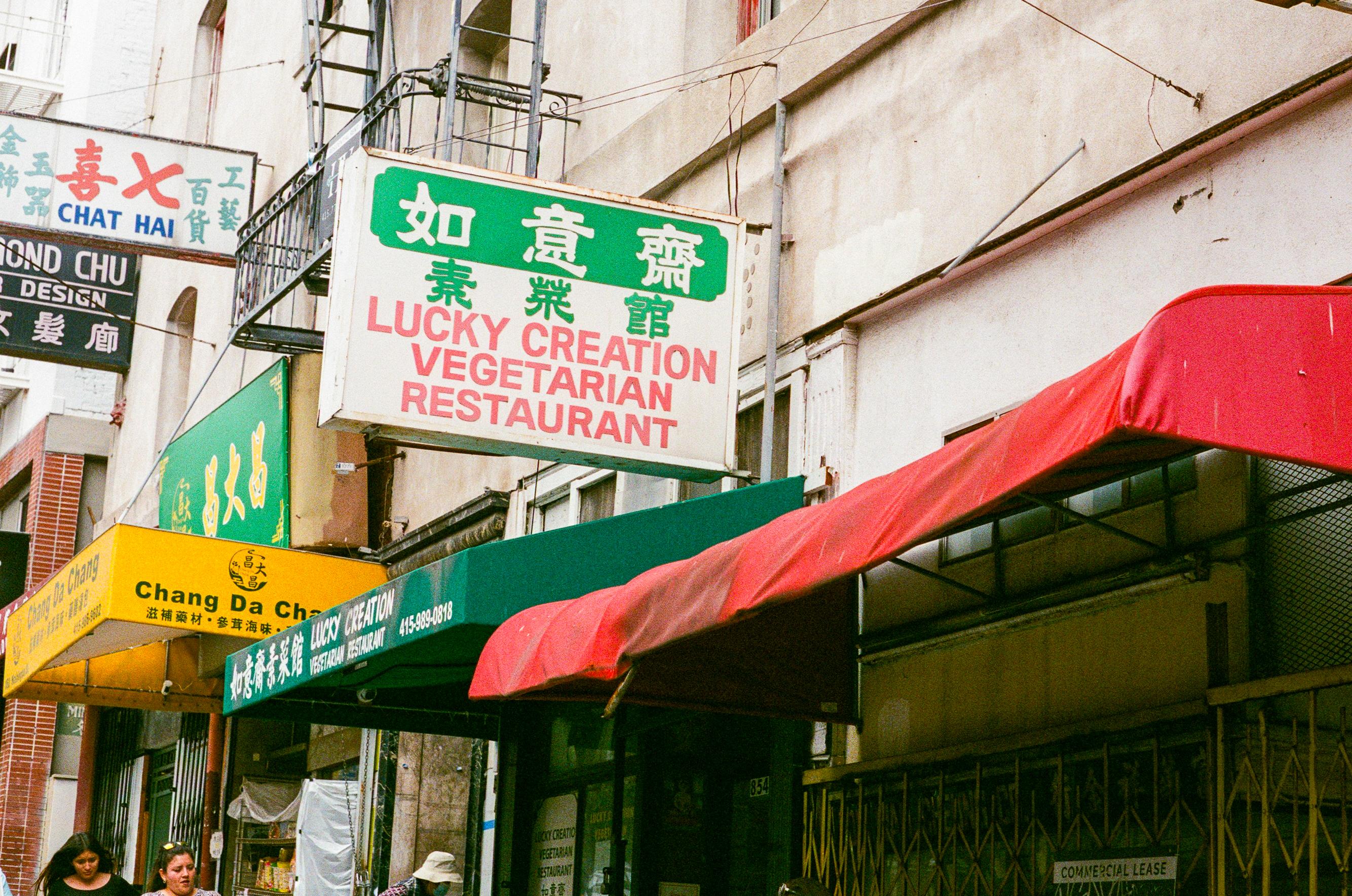 Street view of multiple restaurant signs, including a prominent green and white sign for Lucky Creation Vegetarian Restaurant with a red awning below.