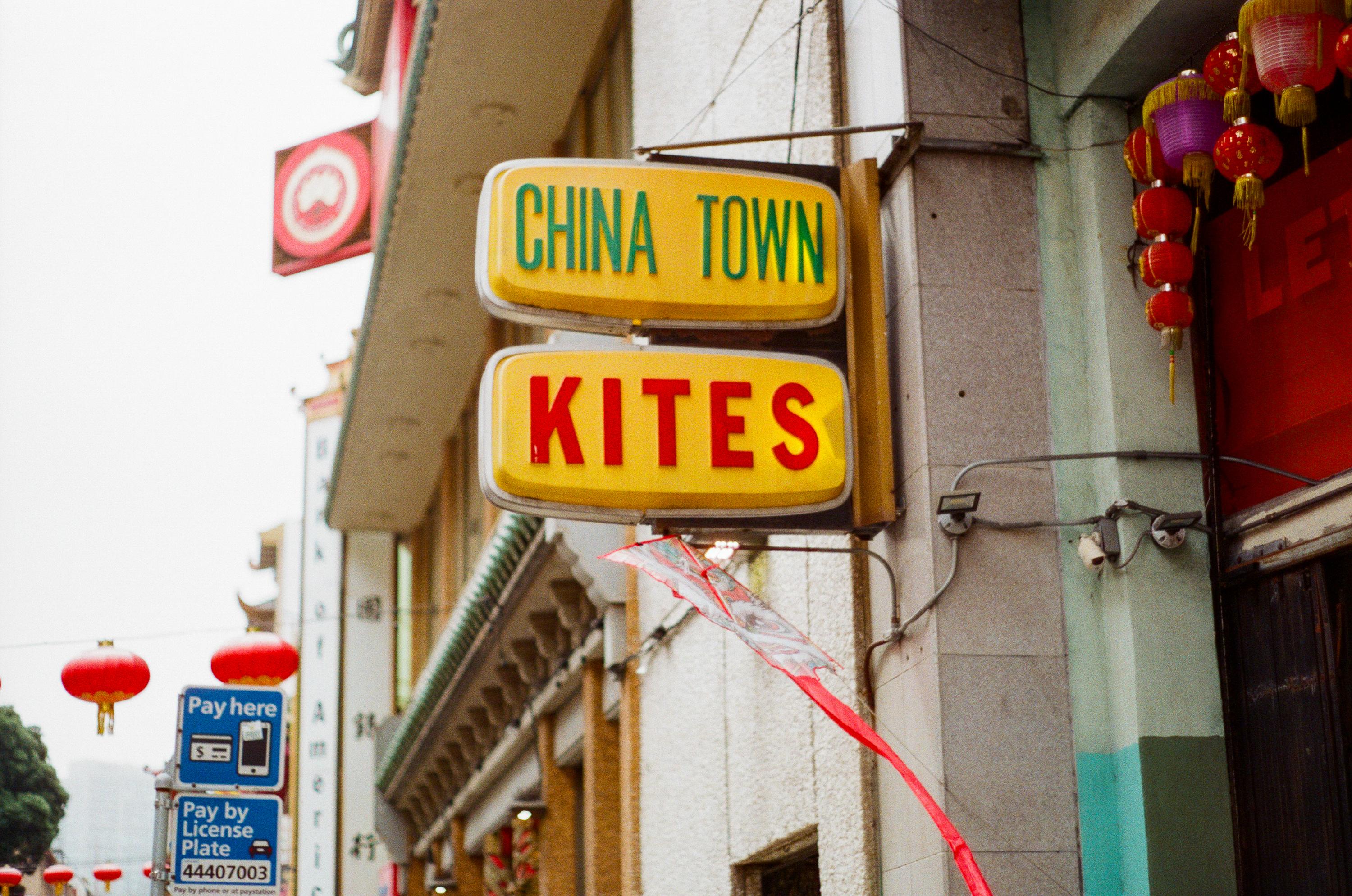 Yellow signs with green and red text reading "CHINA TOWN" and "KITES" hanging on a building facade with red lanterns nearby.