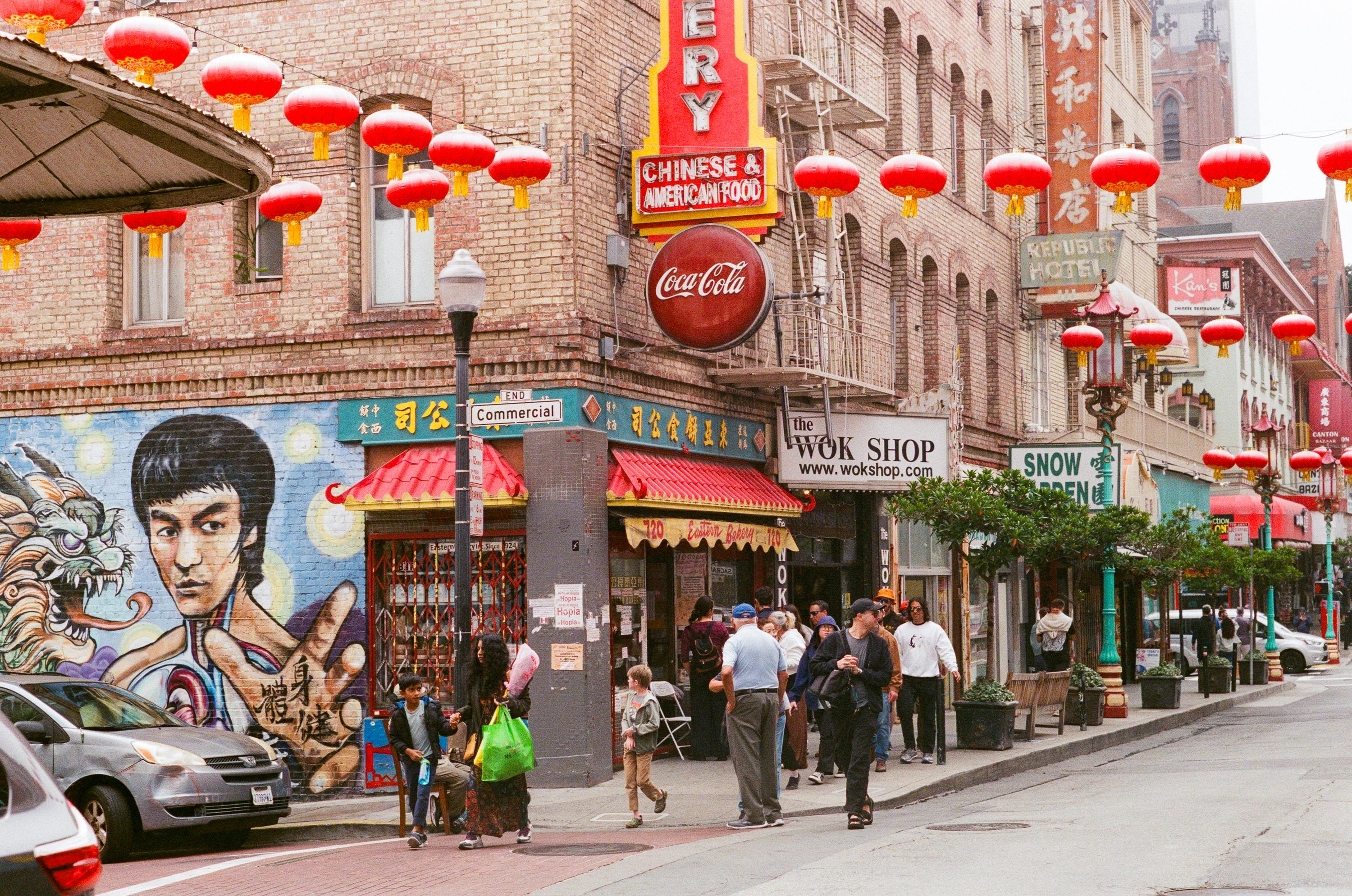 Street scene in Chinatown with red lanterns hanging above, a mural of Bruce Lee, and people walking near restaurants and shops.
