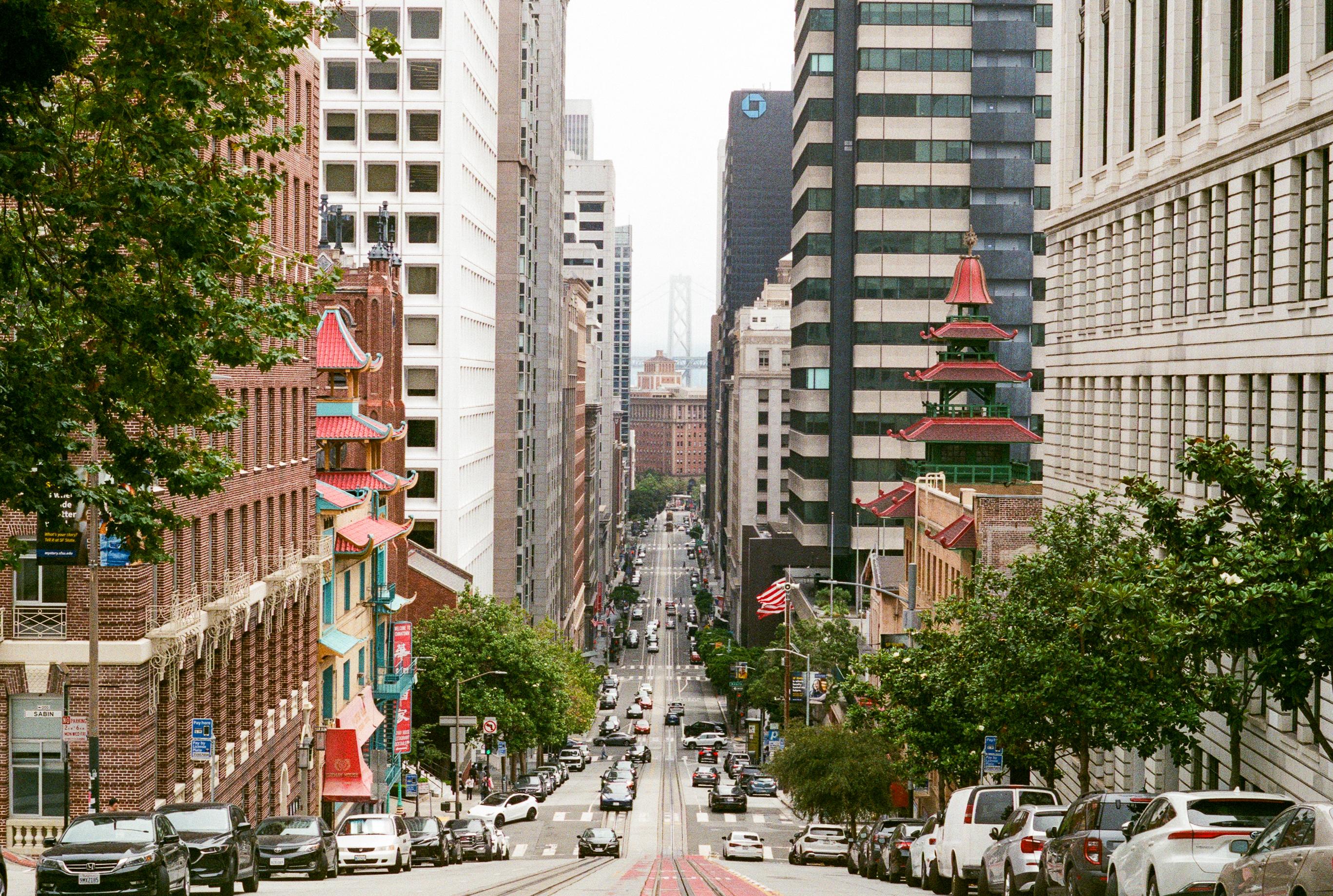 Urban street view with tall buildings, cars parked along the road, and a pagoda-style structure on the right side amidst greenery.