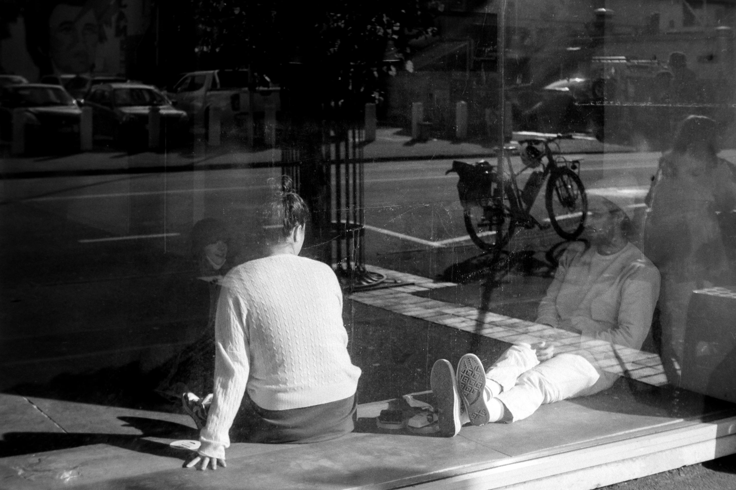 Two people sit inside a glass window display, one facing away, the other reclining. Reflections show a street with parked cars and a bicycle. Black and white photo.